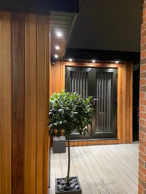 Timber-clad garden room with glazed doors, a planted tree and illuminated decking at night