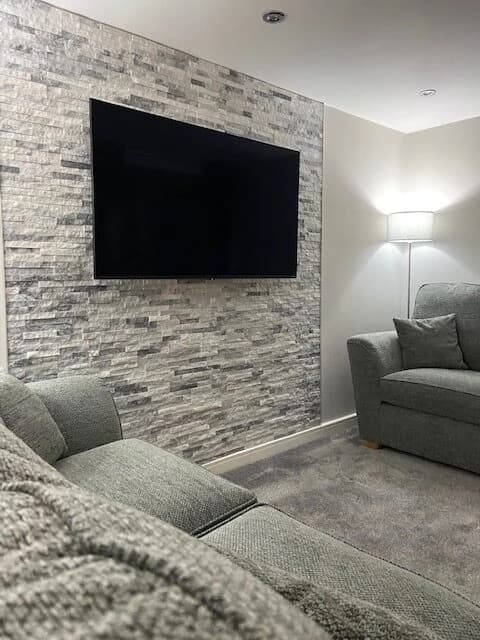 Wall-mounted television on a grey stone-effect feature wall, with a sofa, armchair and floor lamp